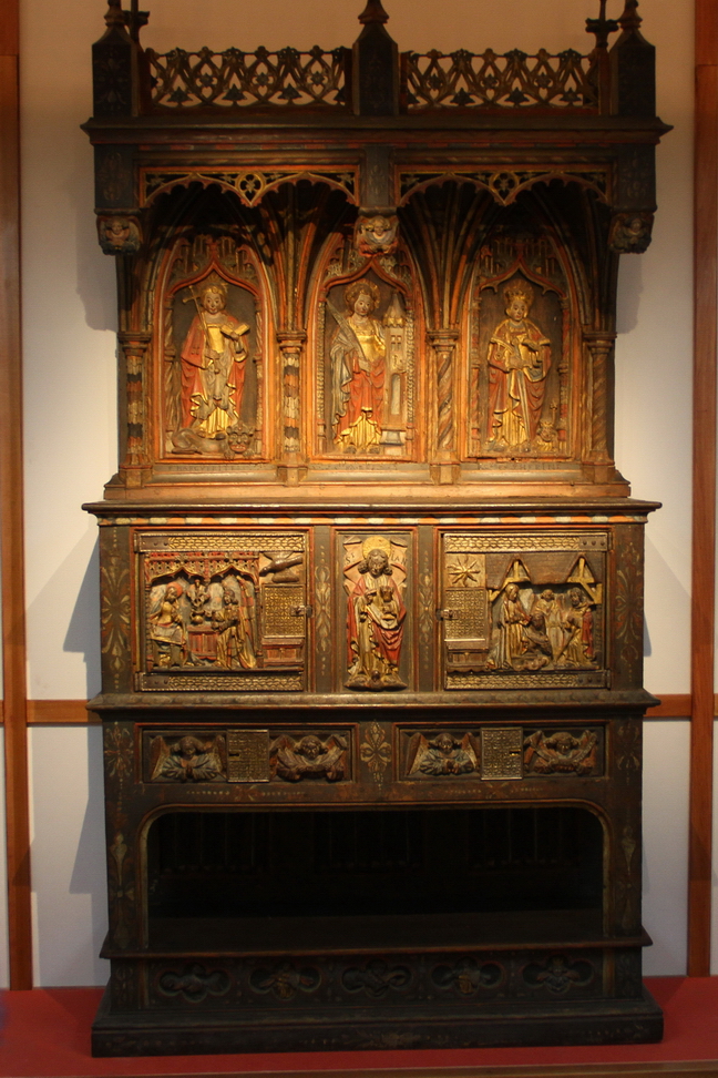 IlCollezionistaDiMeraviglie_055.JPG - Credenza or Dressoir  - Francia settentrionale o Borgogna, seconda metà del XV. - La credenza o dressoir è l'arredo paradigmatico del tardo medioevo e del Rinascimento. Usato per esporre preziose suppellettili, stoviglie e coppe in metallo e in ceramica (il termine francese dressoir deriva da dresser, «esporre», «innalzare»), oltre a manifestare ricchezza e potere fungeva soprattutto da mobile di appoggio nel servizio della tavola (dresser significa anche «apparecchiare, preparare»). Il termine italiano "credenza" fa riferimento all'assaggio preventivo da parte di coppieri e servitori che garantivano così l'assenza di veleni e di sostanze nocive in bevande e cibi destinati alla mensa del padrone di casa e dei suoi ospiti. Il mobile è formato da un corpo centrale a tre ante su piedini, con un'alzata. In alto, sotto il baldacchino, sono raffigurate le sante Margherita, Barbara e Caterina; nel livello inferiore i due sportelli sono decorati con scene dell'Annunciazione e dell'Adorazione dei Magi e separati tra loro dall'immagine di san Giovanni Evangelista. Sui cassetti in basso quattro angeli a mezzo busto reggono cartigli con iscrizioni. L'intaglio ornamentale unisce elementi gotici e rinascimentali: è un esemplare raro, che ha mantenuto in parte la policromia e la doratura originali, anche se alcuni elementi, come le serrature, la zona inferiore e il fastigio sono frutto di restauri ottocenteschi.