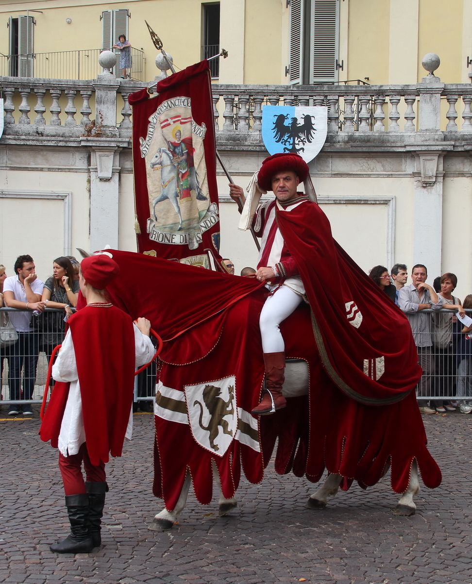 SfilataPalio_018.JPG - SAN SECONDO: la grandezza della vanità terrena "la leggenda dei tre vivi e dei tre morti" - Nel territorio astigiano, a Vezzolano, all’interno del chiostro dell'Abbazia, troviamo una famosa rappresentazione della leggenda "tre vivi e dei tre morti". Leggenda che si diffuse dalla Francia attraverso la Via Francigena e che era tema ricorrente  degli affreschi dei luoghi di culto, poiché accumunava, nella sorpresa e nell'ammonimento, sia i popolani sia gli aristocratici Il Rione San Secondo rappresenta, l'iconografia "dei “tre vivi e dei tre morti" attraverso scenografici quadri viventi: i “tre vivi” compaiono nelle vesti di tre giovani signori, dediti  alla nobile arte della  caccia con il falcone, allora diffusa presso l’alta nobiltà feudale. I tre giovani, durante la battuta di caccia, s'imbattono in “tre morti”, che dichiarano: "Io fui Papa"; "Io fui Cardinale"; "Io fui Notaio apostolico".  Poco distante si scorge un eremita che mostra una pergamena in cui è riportato un ammonimento "Voi sarete come noi: potere, onore, ricchezza sono vani". I cavalieri fuggono atterriti, ma poco dopo compare loro una croce e comprendono di aver ricevuto un segno divino che li esorta a rifuggire le vanità terrene  e a rivolgere i loro pensieri a Dio.