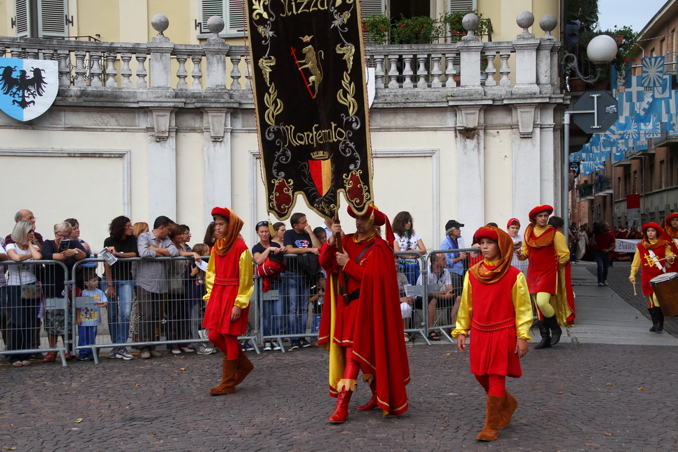 SfilataPalio_044.JPG - NIZZA MONFERRATO: Kalenda Maya - Kalenda  Maya“…non fragor d’armi ne esce, né gridar di contese; la guerra che ogni primavera torna a insanguinare… non trova eco nei versi di coloro che per primi poetarono nella nostra lingua materna”: così canta Raimbaud de Vacqueiras, nato in Provenza attorno al 1165, figlio di un cavaliere spiantato e stravagante. Giovanissimo apprende l’arte dei trovatori e diventa joglar (giullare, menestrello) alla corte di Guglielmo di Baus, Principe d’Orange. Accanto all’arte del comporre e raccontare in musica, affina l’arte delle armi, nella quale si afferma al pari del canto. Ma a seguito delle violenze che sconvolgono la Provenza Raimbaud, come altri joglar, abbandona la sua terra e percorre le strade del Nord Italia fino a stabilirsi alla corte del Marchese Bonifacio del Monferrato (1192). Al suo fianco combatte contro la Città di Asti, nella campagna di Sicilia e nella Quarta Crociata. Durante il suo soggiorno alla corte del Monferrato si innamora di Beatrice, figlia naturale del Marchese, poi sposa di Enrico del Carretto: per la donna amata, che Raimbaud chiama “Bel Cavaller”,  compone le sue canzoni. Joglar, Trobadour, Minnesänger erano maestri di festa e di gioia presso le corti medievali e Raimbaud de Vacqueiras lo fu in modo assoluto ed innovativo alla corte di Bonifacio di Monferrato. Le sue composizioni poetiche hanno resistito all’usura dei secoli e sono note ancora oggi. Fra tutte eccelle il Kalenda Maya. Alla corte di Bonifacio la musica, il canto e la prosa allietavano il tempo libero e la festa: con la primavera si rinnovavano gioia, vitalità e amori dopo i rigori dell’inverno. I musici e le giovani; i cantori, le dame, i cavalieri intrecciavano i loro corpi in balli gioiosi fra i fiori dei giardini e le rose di maggio. La frutta più bella e la cacciagione. ….E l’amore che esplode tra i giovani a primavera. L’amor cortese è l’amor profano. Il trovatore canta per l’onore del Marchese e Signore delle terre che vanno dal mare fino a Casale, da Lanerio a Chivasso per tutta la pianura del Tanaro e del Po. E Nizza, non ancora città, ma terra di Lanerio, Belmonte e Quinciano si appresta a diventare monferrina.