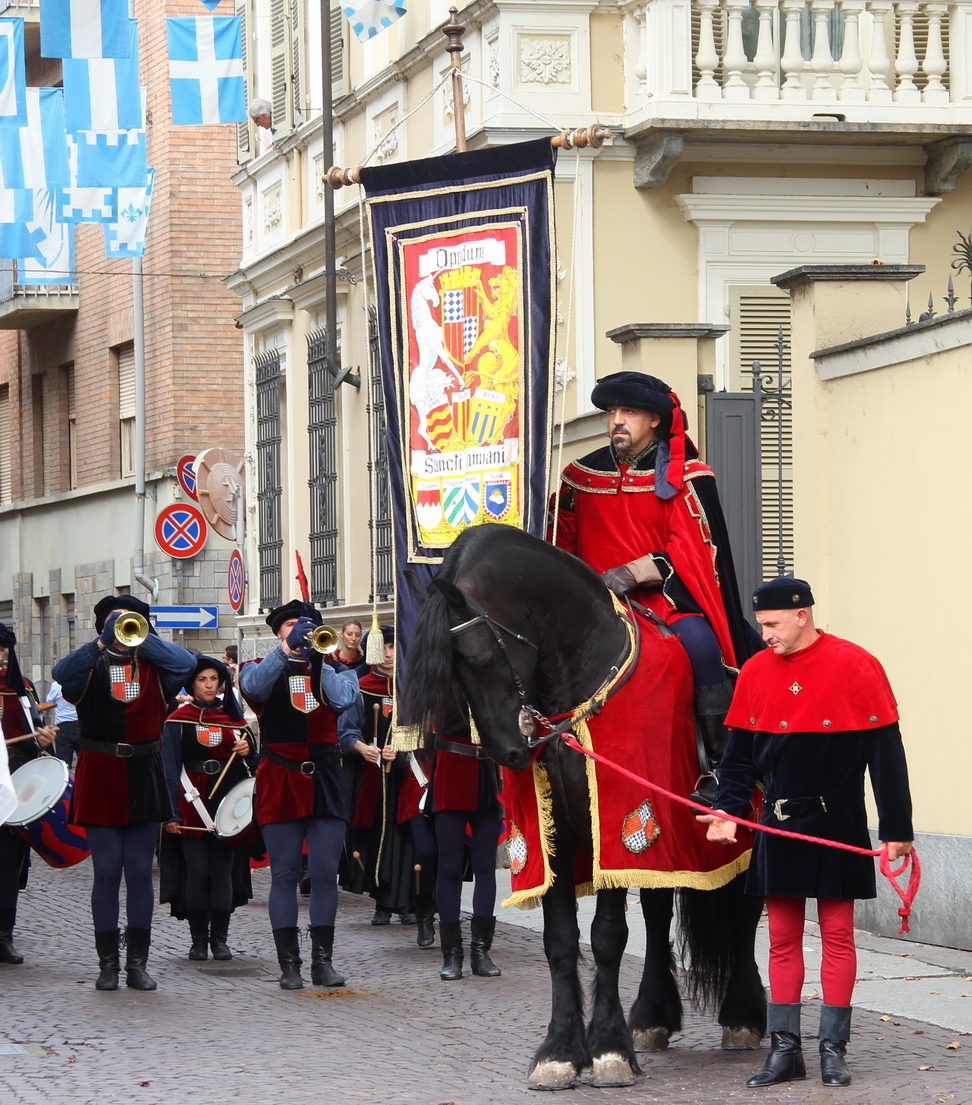 SfilataPalio_072.JPG - SAN DAMIANO: "Questi Lombardi cani" - Gli usurai astesi e la dinastia Scarampi - I  “Questi Lombardi cani”: gli usurai astesi e la dinastia ScarampiLo sviluppo economico del basso medioevo modificò il commercio del denaro, attività condannata dalla Chiesa e pertanto a lungo demandata agli Ebrei. Il prestito era però indispensabile per sostenere lo sviluppo economico e riprese vigore dapprima grazie all’attività dei Templari, che  raccoglievano e gestivano  denaro per conto del papa per finanziare le crociate e in seguito innovarono la pratica bancaria in Occidente. Accanto ai monaci guerrieri il prestito era praticato da altre categorie di operatori,  tra questi i “lombardi”, prestatori originari dell’Italia centro-settentrionale. Gli Astigiani furono tra i più attivi rappresentati di questa categoria, che talora era oggetto dello stigma sociale, come ben illustra la  novella del Decamerone nella quale sono definiti ‘Lombardi cani’.Ogerio Alfieri nella sua cronaca scrive che gli Astigiani incominciarono ad esercitare su vasta scala il commercio del denaro  nel 1226: si trattava di un’evoluzione rispetto all’attività di cambiatori che praticavano da tempo nelle grandi fiere e nei mercati europei.Gli Astigiani acquistavano a Genova le merci provenienti dall’Oriente destinate alle fiere dell’Europa centro-settentrionale: la documentazione dei notai genovesi, infatti, a partire dal 1190 attesta la presenza dei mercanti astesi alle fiere di Champagne, ove vendevano pepe, spezie, allume, sete, argenterie e gioielli e acquistavano, per rivenderli al di qua delle Alpi, lane e panni di Douai, Tournay, Arras, stanforti d’Inghilterra, Fiandra e Artois. Come si è ricordato, l’intenso movimento di denaro e la capacità di gestire le pratiche di cambio favorirono la trasformazione dei mercanti/cambiavalute in prestatori su pegno, dapprima individualmente, ma ben presto in forma associata nelle cosiddette casane.  Il commercio del denaro rese Asti una delle città più ricche del nord Italia: le famiglie più abbienti acquistarono feudi e castelli, fecero proprio uno stile di vita simile a quello della nobiltà, strinsero potenti amicizie; in città sorsero chiese, torri e palazzi sontuosamente arredati, il lusso venne ostentato. Come tramandano i cronisti, le donne del patriziato astese sfoggiavano lussuose vesti adorne di decorazioni in oro e argento, con pellicce e preziosi gioielli ornati di perle e pietre preziose. Tra le famiglie nobili astesi, la dinastia degli Scarampi praticò il prestito su pegno in vari mercati europei, prestando denari a principi, re, nobili e alti prelati; furono però accusati di usura e vennero espulsi da molte città e regni, al punto che nelle Fiandre era da ritenersi una grave ingiuria definire qualcuno “Scarampi”, denominazione intesa come sinonimo di usuraio. Il corteo rosso blu rievoca l’attività di uno degli esponenti della dinastia astese Daniele Scarampi, morto nel 1446 e sepolto nella chiesa della Maddalena di Asti. Ancor oggi la sua lastra tombale, custodita a San Damiano, è testimonianza dell’antico legame della nobile famiglia con questo territorio.