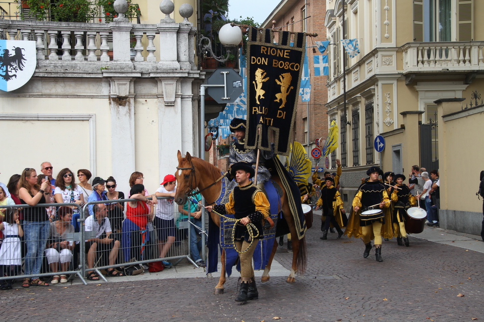 SfilataPalio_082.JPG - DON BOSCO: Il "Districtus civitatis" - Allegoria di un dominio territoriale - Il “Distirictus  Civitatis”, allegoria di un dominio territorialeA partire dal XIV secolo le grandi famiglie magnatizie astigiane iniziarono sistematicamente ad investire i ricchi proventi dell’attività bancaria secondo strategie di insignorimento territoriale, acquistando dal Comune la giurisdizione di numerosi villaggi e castelli ad esso appartenenti. Tali iniziative, benché formalmente non mettessero in discussione l’integrità della “patria astese” e i diritti della dominante, di fatto sottraevano alla “res publica” quote importanti di giurisdizione e alle casse comunali rilevanti introiti ed emolumenti ad essa collegati. Per ovviare almeno in parte ad un fenomeno che sembrava dolorosamente inarrestabile, Asti, nel corso del Trecento, elaborò e definì il concetto di “Districtus Civitatis”: una parte del territorio che, sulla base di rigide norme statutarie e legali, doveva rimanere sotto l’inalienabile e diretto dominio della Città.Tale ripartizione assunse definitiva forma giuridica sotto la signoria di Gian Galeazzo Visconti: essa comprendeva i villaggi a corona della Città che ancora oggi costituiscono il suo territorio comunale, nonché una serie di comuni confinanti per un raggio di circa dieci chilometri. Tutte le terre del districtus dovevano essere governate da un podestà nominato annualmente da Asti e scelto in seno al Consiglio Generale della Città; la diretta dipendenza dalla “capitale” prevedeva inoltre una serie di oneri fiscali, tributari e legislativi. E' del 1385 la prima completa elencazione delle "ville" che costituivano il Distretto. Esse erano le attuali frazioni di Serravalle, Sessant, Mombonino, Variglie, Castiglione, Quarto, Montemarzo, San Marzanotto, Bellangero e i comuni di Cinaglio, Camerano, Antignano, Celle Enomondo, Tigliole, Baldichieri, Villafranca, Cantarana, Azzano, Mongardino, Vigliano, Isola d’Asti, Montegrosso d’Asti, Portacomaro, Scurzolengo e Castell’Alfero. Con poche variazioni territoriali, il dominio sul Distretto cittadino,  una vera e propria "area metropolitana" ante litteram,  fu a lungo gelosamente e caparbiamente difeso da Asti, che intravvedeva in esso l'irrinunciabile baluardo delle sue residue libertà comunali ed un importante presupposto di prosperità economica e di prestigio urbano. Del “Districtus Civitatis” ebbero ragione solo le politiche accentratrici ed assolutistiche dei Savoia, che a partire dal 1618 ne  provocarono il progressivo smantellamento  .