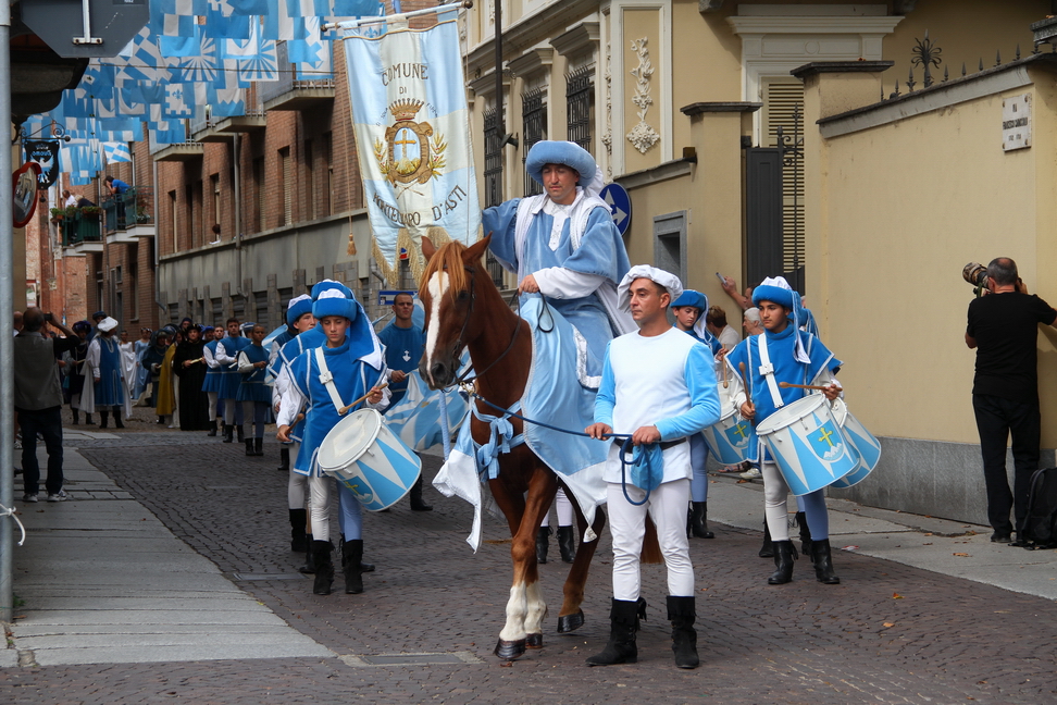 SfilataPalio_116.JPG - MONTECHIARO - Gli abitanti di Montechiaro giurano fedeltà a Valentina Visconti - Nel 1387 Gian Galeazzo Visconti, dal 1379 Signore di Asti, concedeva la città ed il territorio da essa dipendente in dote alla figlia Valentina per il suo matrimonio con il Duca Ludovico di Touraine, fratello del Re di Francia Carlo VI.Il perfezionamento di un atto politico di questa portata necessitava di una complessa procedura insieme giuridica e simbolica, che prevedeva, tra l’altro, il giuramento di fedeltà dei cittadini a Valentina.Anche gli abitanti di Montechiaro, paese sul quale il Comune di Asti esercitava da secoli il proprio controllo e destinato a passare sotto la nuova dominante, dovettero dichiarare esplicitamente la loro fedeltà ai nuovi signori: il 26 maggio 1387 a Montechiaro nella chiesa di Santa Maria Maddalena i cives si presentarono davanti ai delegati ducali, Bertramo Guasco per Valentina e Catelano de Christianis per Ludovico, e giurarono fedeltà e omaggio a Valentina e al suo sposo Ludovico nelle mani del notaio Jean Sicard, toccando con la mano il Santo Vangelo.Il documento che attesta questo episodio riporta tutti i nomi degli abitanti coinvolti nella cerimonia solenne: giurarono soltanto i cives a pieno titolo, ovvero i maschi di età superiore ai quattordici anni, mentre furono esclusi i religiosi e i nullatenenti. Furono parimenti escluse le donne, nobili, borghesi o popolane,Nella ricostruzione dell’episodio  il corteo biancoceleste vuole rappresentare il momento del giuramento facendo sfilare i protagonisti, ovvero il Notaro, i delegati Ducali e i cives, ma anche gli esclusi: i religiosi che recano i Sacri Testi, i nullatenenti  che seguono il corteo rumoreggiando e chiedendo la carità, dame, damigelle, borghesi e popolane che accompagnano i propri congiunti ed infine i portacolori.