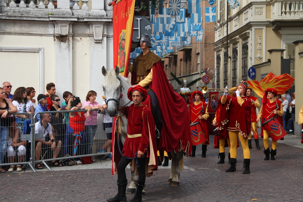 SfilataPalio_120.JPG - SAN PAOLO - La processione della domenica in occasione delle festività patronali nella Asti Medioevale - Nella Asti medioevale due ricorrenze, una volta all’anno, coinvolgevano ed entusiasmavano la popolazione, sia in città sia nel  contado, rompendo la monotonia della vita di tutti i giorni: la festa patronale e la corsa del palio. La festa patronale affiancava alla componente religiosa anche significati politici, con aspetti di chiara valenza simbolica: tra questi il cero, un omaggio che già nel XII secolo il Comune imponeva a comunità e signori sottoposti al suo controllo, tenuti a partecipare alla cerimonia in onore del Patrono di Asti .Anticamente la festa patronale veniva celebrata nel giorno anniversario del martirio di San Secondo, il 30 marzo; poiché, però, questa data sovente cadeva durante la Settimana Santa o in Quaresima, la festività venne spostata al giovedì che seguiva la prima domenica in Albis, cioè successiva alla Pasqua. Le funzioni della domenica antecedente la festa patronale erano riservate al Capitolo della Collegiata di San Secondo. Al mattino si cantava Messa solenne con assistenza pontificale e con intervento ufficiale delle autorità. Alla sera veniva portato in processione il busto d’argento del Patrono, seguito dalle più eminenti personalità e dai giovani paggi prescelti per la corsa al Palio, che avrebbe coronato le celebrazioni patronali Il Rione San Paolo rappresenta questa processione in onore del Patrono: accompagnano  l’effige del Santo i componenti del clero cittadino, le autorità del Comune, nobili e nobildonne delle famiglie astigiane, i rappresentati di comunità e signori sottoposti al controllo di Asti che recano i ceri votivi, mercanti, banchieri, uomini di legge e una simbolica rappresentanza dei giovani paggi che correranno il Palio.