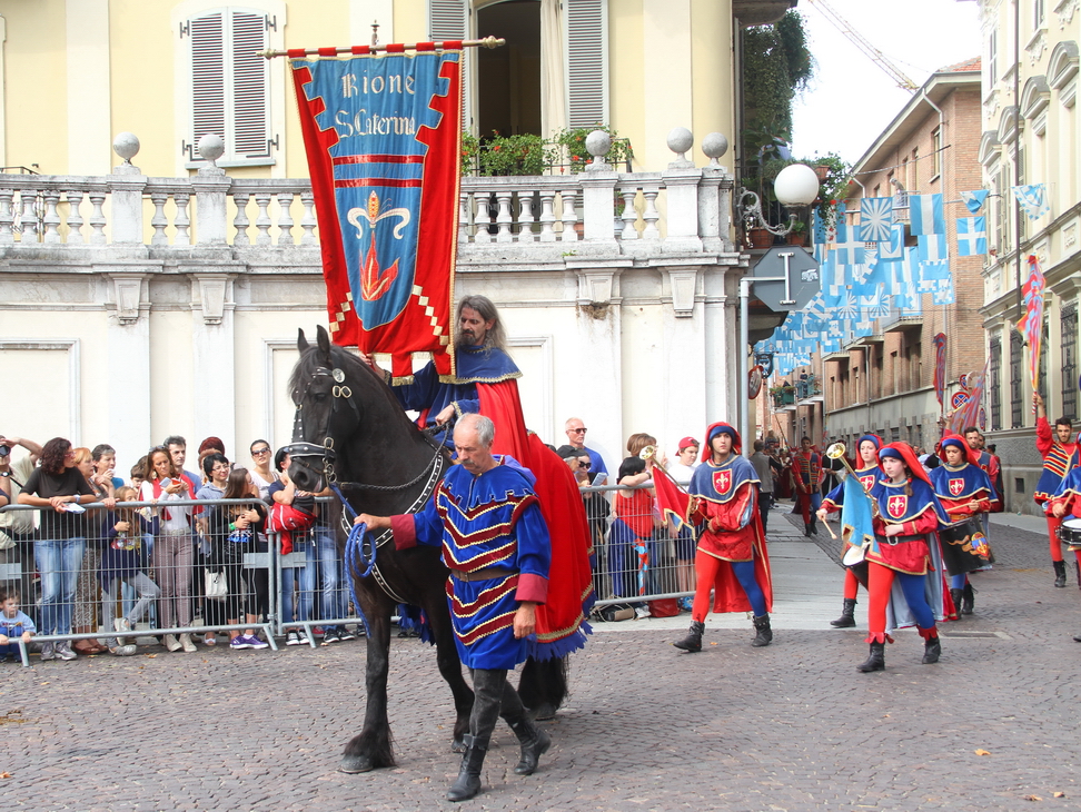 SfilataPalio_161.JPG - SANTA CATERINA - La “Legenda aurea”: Santa Caterina giovane martire cristiana - La tradizione culturale medievale ci ha consegnato una monumentale opera agiografica, la Legenda aurea di Jacopo da Varazze (1228 c.a./1298), frate domenicano che fu anche priore dell’ordine ad Asti nella seconda metà del XIII secolo.Nella Legenda aurea vengono narrate le vite esemplari di cento ottantadue santi, modelli di vita cristiana: tra questi anche Santa Caterina di Alessandria d’Egitto, alla quale è dedicata la chiesa del rione.La giovane martire ha avuto  grande importanza nella tradizione cristiana - classica e medievale - ,  nella vita di numerosi ordini religiosi, nella storia degli studi filosofici e teologici, nelle tradizioni popolari e nell’iconografia religiosa, come dimostrano le numerosissime raffigurazioni della Santa  nel corso dei secoli. Secondo il racconto di Jacopo da Varazze, l’egiziana Caterina, figlia di re Costo, fu istruita fin dall'infanzia nelle arti liberali. Nel 305 Caterina si presentò a palazzo ad Alessandria durante i grandi festeggiamenti organizzati dall’imperatore romano Massenzio, nel corso dei quali si celebravano feste pagane con sacrifici di animali. Caterina rifiutò i sacrifici e chiese all'imperatore di riconoscere Gesù Cristo come redentore dell'umanità, argomentando la sua tesi con profondità filosofica.L'imperatore, che, secondo la Legenda Aurea, sarebbe stato colpito sia dalla bellezza sia dalla cultura della giovane, convocò un gruppo di retori affinché la convincessero ad onorare gli dei. Tuttavia, grazie alla profondità della fede di Caterina, non riuscirono nell’intento, anzi essi stessi furono prontamente convertiti al Cristianesimo per merito della sua eloquenza .L'imperatore ordinò la condanna a morte di tutti i retori e dopo l'ennesimo rifiuto di Caterina la condannò a morire anch'essa su una ruota dentata. Tuttavia lo strumento di tortura si ruppe e Massenzio fu obbligato a far decapitare la santa.Il suo di Caterina venne poi trasportato dagli angeli fino al Sinai, la Montagna di Mosè dove si ergeva un famoso monastero in seguito dedicato alla martire.Il corteo rosso-azzurro, rifacendosi alla versione della Legenda Aurea, ripercorre i momenti fondamentali della vita della Santa: la conversione, la disputa con i retori, il martirio della ruota e infine la morte per decapitazione. 