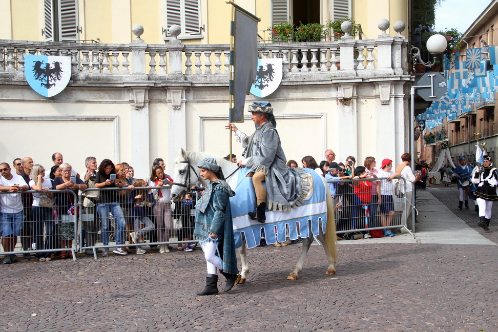 SfilataPalio_175.JPG - VIATOSTO - Tra cielo e terra: simbologia e mistica degli alberi nel Medioevo - Alberi e piante hanno da sempre affascinato l’uomo. Nel Medioevo la simbologia collegata al culto degli alberi assunse significati religiosi cristiani, che si sovrapposero a miti e leggende antiche di matrice celtica. Il Borgo Viatosto rende omaggio al patrimonio naturale che ancora oggi ne costituisce la preziosa cornice, rappresentando in un’allegoria alcuni alberi e arbusti che si caratterizzavano per il loro particolare significato: la rosa, emblema di purezza e intimamente collegata al culto mariano; il noce, albero collegato a culti stregoneschi che sfruttavano le supposte proprietà divinatorie dei suoi frutti; l’olmo, sotto le cui fronde in Francia veniva amministrata la giustizia; il tasso o “albero della guerra” e della caccia, dal legno flessibile, particolarmente adatto per la fabbricazione degli archi.Non possono mancare i tre elementi simbolici presenti all’interno della chiesa romanica di Viatosto: il grano, le cui spighe costituiscono una premessa di benessere e nutrimento per le generazioni; la quercia, albero della legna, i cui frutti costituiscono un prezioso nutrimento per molte specie animali, presente sulla volta in una sorta di immensa ghirlanda; l’albero delle ciliegie: queste ultime sono rappresentate nella pala d’altare sulla navata sinistra dedicata alla Madonna, e costituiscono il simbolo della passione di Cristo, rinsaldando il legame tra natura e religiosità che costituisce l’elemento caratterizzante della storia del Borgo.