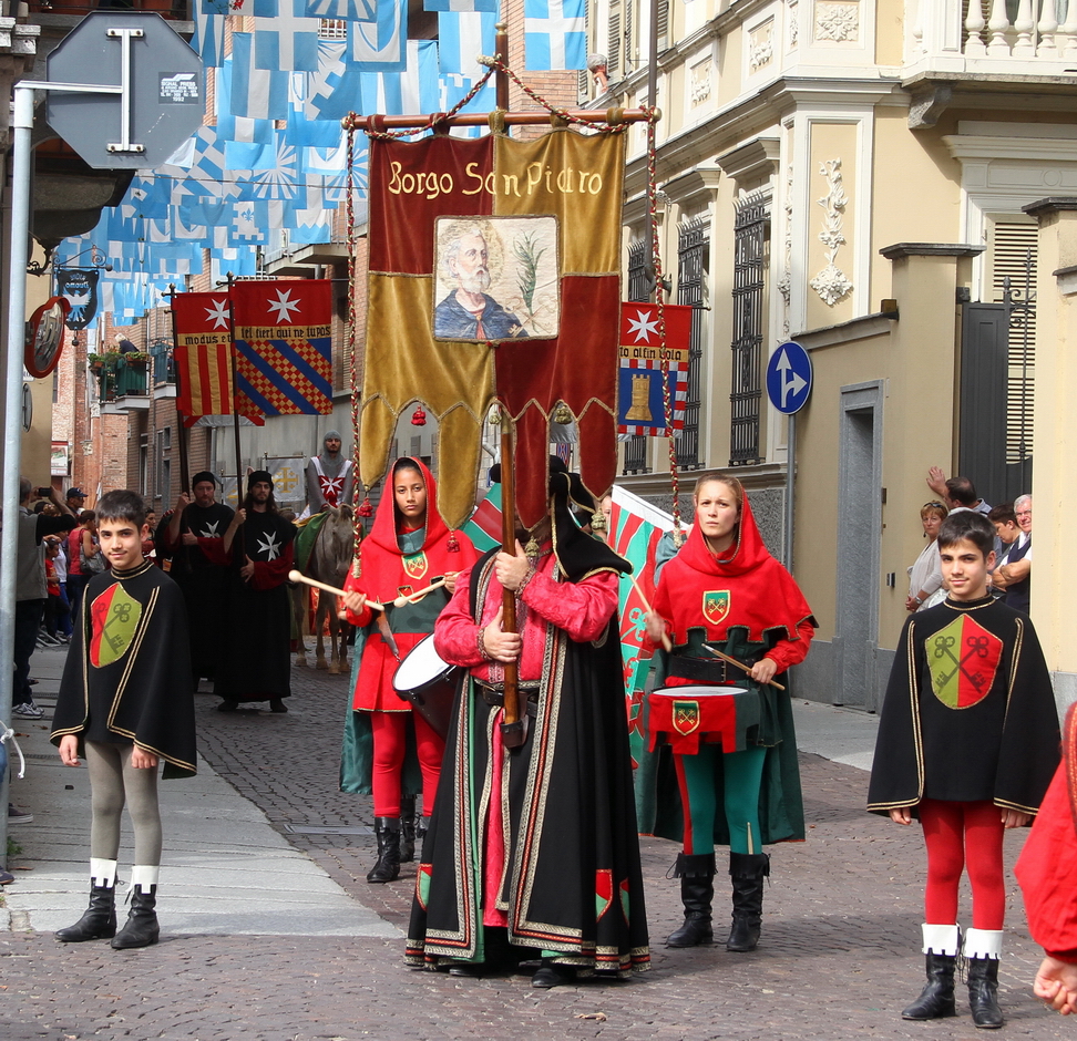 SfilataPalio_193.JPG - SAN PIETRO - In cammino verso Gerusalemme tra fede e meraviglia - La ricerca storiografica più recente ha modificato l’immagine tradizionale dell’uomo medievale chiuso entro le mura della città o del monastero, dimostrando invece come fossero frequenti, nonostante le difficoltà, i viaggi di mercanti, monaci, cavalieri e pellegrini che per motivi politici, economici, religiosi e militarisi ponevano in cammino.Posta al centro del sistema viario della pianura padana, da sempre snodo importante della via Francigena, Asti vide confermato il suo ruolo centrale rispetto agli itinerari diretti verso i luoghi sacri con la bolla del 1113, con la quale papa Pasquale II riconobbe ufficialmente l’Ordine dei Cavalieri dell’ Ospedale di Gerusalemme: in essa Asti è citata, unica città dell’entroterra, insieme a sei porti marittimi d’imbarco per l’Oriente. Fu proprio lungo l’asse dell’antica via Fulvia che ad Asti si insediarono i Gerosolimitani e sorse così San Pietro in Consavia, che anche nella forma architettonica richiama la rotonda del Santo Sepolcro con precisi riferimenti a Gerusalemme.Molti furono gli astigiani che, entrati nell’ordine Gerosolimitano, partirono desiderosi di conoscere mondi nuovi, spinti dal forte richiamo della difesa dei luoghi santi e guidati anche da uno spirito errabondo ed avventuroso. Il viaggio, quasi sempre lungo e difficile, diventò una prova di vita superabile con la fede, paradigma del passaggio dal mondo del peccato alla salvezza attraverso la penitenza e il riscatto. Meta reale e spirituale del cammino era Gerusalemme, posta in molte carte geografiche al centro dell’ Universo tra il Paradiso e l’Inferno. A testimonianza di questi viaggi, i racconti di cavalieri e pellegrini che si avventuravano verso l’ ignoto narravano spesso di forme di vita meravigliose e mostruose al confine fra l’umano e il sovraumano: l’ uomo-bestia, il serpente crestato, il basilisco, le fanciulle-fiore, le sirene, che compaiono anche nelle decorazioni di San Pietro in Consavia, a simboleggiare le paure, le illusioni e le speranze di coloro che viaggiavano.  