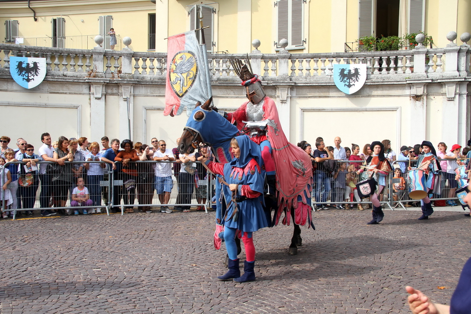 SfilataPalio_216.JPG - SANTA MARIA NUOVA - San Biagio e la festa della Candelora - Nel Borgo di santa Maria Nuova fin dal medioevo si diffuse il culto di San Biagio;  nel XV secolo il santo  divenne compatrono della chiesa insieme alla beata Vergine ed alcuni resti dello stesso sono conservati nell'altare maggiore. Durante la funzione religiosa per la festa, oltre alle candele, venivano benedetti dei pani  consumati dopo la processione, gli avanzi venivano conservati ed utilizzati come curativi delle affezioni della gola ed i ceri venivano conservati nelle abitazioni dei fedeli per essere riutilizzati in caso di necessità. In epoca medievale nel borgo di Santa Maria Nuova  era particolarmente venerato San Biagio, le cui reliquie erano conservate nell'altare maggiore della chiesa. Nel XV secolo, quando fu costruito il nuovo edificio sacro, San Biagio ne divenne compatrono insieme alla beata Vergine della Purificazione. I Canonici Lateranensi, cui la chiesa fu affidata a partire dal 1472, commissionarono a Gandolfino da Roreto due affreschi ai lati del portale d'ingresso, raffiguranti il protettore della Congregazione,  Sant'Agostino e San Biagio. Quest'ultimo era un medico armeno vissuto tra il III e IV secolo a Sebaste in Asia Minore, perseguitato e decapitato dai romani perché rifiutò di rinnegare il Cristianesimo. Nella sua qualità di medico, viene invocato dai fedeli in particolare per la guarigione dalle malattie della gola. È anche protettore dei cardatori di lana, degli animali e delle attività agricole.La festa di San Biagio ricorre il 3 febbraio e segue quindi quella della Purificazione della Vergine, cui la chiesa è dedicata, detta anche ”Candelora” per la tradizionale benedizione delle candele tipica di tale Festività. Sono ricorrenze che rimandano ad antiche tradizioni pagane collegate alla fine dell'inverno: secondo i celti, le grandi piogge di febbraio erano segni di purificazione della terra e questa data segnava il passaggio tra l’inverno e la primavera, tra il buio e il freddo e il risveglio della luce. Le manifestazioni cristiane si sono sovrapposte ai riti pagani e le feste della Purificazione della Vergine e di San Biagio  coincidono con l’offerta di candele benedette della "Candelora". Durante la celebrazione in onore del Santo, oltre alle candele, venivano benedetti dei pani dolci o salati, che venivano consumati dopo la funzione.  Il pane che avanzava veniva conservato ed utilizzato come cura delle affezioni della gola. I ceri usati nella processione erano conservati nelle abitazioni dei fedeli per garantirsi l’intercessione di San Biagio durante calamità meteorologiche, nell'assistenza di un famigliare gravemente ammalato o nel caso di epidemie. Il corteo storico, preceduto da vessillifero, sbandieratori e musici,  è aperto dal priore di Santa Maria Nuova che reca le caratteristiche candele incrociate per la benedizione della gola. Lo accompagnano due religiosi: uno mostra le reliquie del Santo, mentre l'altro porta il pane benedetto. Segue il "paliotto" di San Biagio protettore della  corporazione dei lanaioli, accompagnato da esponenti della famiglia dei Lupi - drappieri che esercitavano la loro attività commerciale presso il borgo di Santa Maria Nuova - i quali portano le candele tipiche della "Candelora".