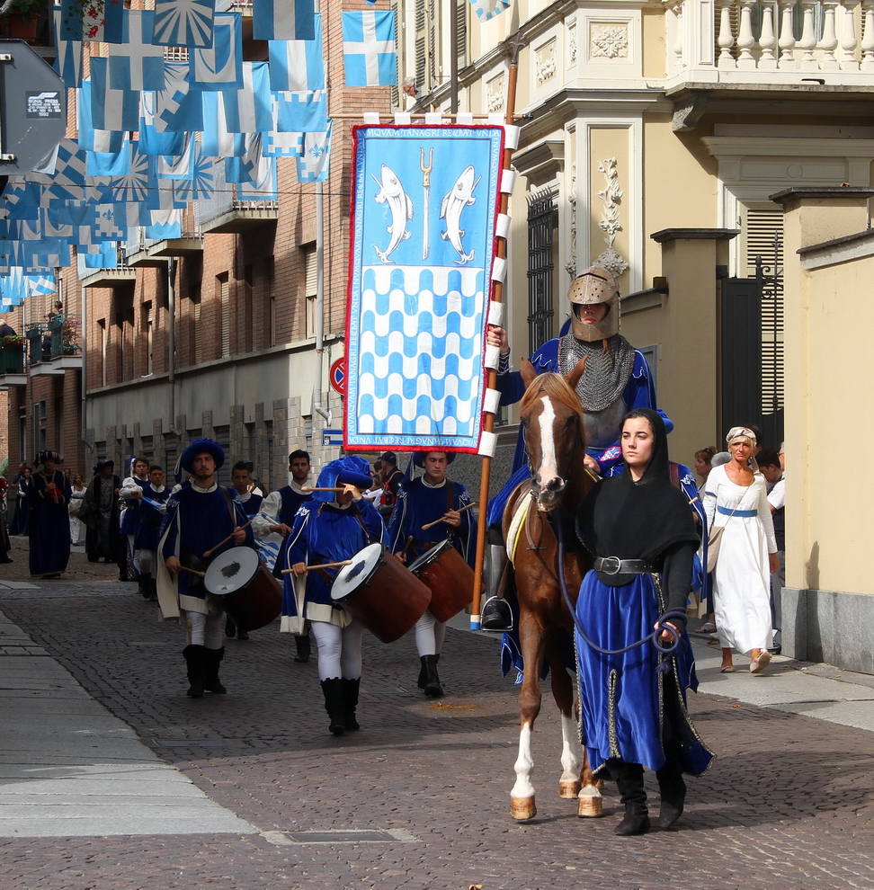 SfilataPalio_225.JPG - BORGO TANARO TRINCERE TORRAZZO - La vita sul fiume: dagli affari alle punizioni. La nascita della Società del Moleggio - Travagliata da continue guerre e lotte civili, l’economia astigiana nella seconda metà del XIV secolo era pesantemente compromessa. Luigi d’Orleans, signore di Asti dal 1387, decise di stimolare la ripresa economica con un’iniziativa imprenditoriale di grande rilievo. “…Si aqua fluminis Trevezie in civitatem nostram prolictam duceretur…..propter molendine, batanderia, parateria ut recas et alia artificia ibidem costruenda et edificanda…”: così si legge nelle Lettere Patenti  del 23 ottobre 1397, documento  mediante il quale  il Signore d’Orleans sancì   la nascita della Società del Moleggio, cui assegnava il monopolio   sulla macinazione cerealicola nell’astigiano. La somma di 10.560 genoini, pari a 46.464 lire astesi dell’epoca, venne investita metà da sedici famiglie nobili astigiane e metà dallo stesso Luigi d’Orleans per la realizzazione delle bealere e la costruzione di nove mulini, di cui quattro molini natanti sul fiume Tanaro. Tra le famiglie nobili astigiane associate all’iniziativa comparivano i Curia, i Solaro, i Laiolo, gli Asinari, i Riccio ed altri ancora. Questa impresa doveva ripristinare l’attività molitoria,   allora in crisi, ma al contempo creare nuovi mulini, battitoi, paratoi e segherie per ridare prosperità e benessere alla città e incrementare il commercio sul fiume.Grazie a questo investimento il   Tanaro assumeva una centralità nuova nella    vita economica e commerciale della città. Gli approdi fluviali tornarono ad essere luoghi di commercio, ove cercare stoffe, spezie e prodotti alimentari, vi trovarono spazio il mercato del pesce, l’attività delle lavandaie e i frutti della fatica dei contadini. Ma il fiume poteva essere anche teatro dell’amministrazione della giustizia:  negli ordinati comunali di fine XIV secolo si legge che la pena per i bestemmiatori consisteva nell’essere  calati con una corda dal ponte del Tanaro nelle acque del fiume sino a che non ne venivano completamente sommersi.